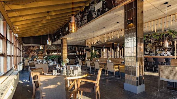 Un amplio comedor interior con mesas y sillas de madera, un techo alto con vigas a la vista y una zona de bar con taburetes.