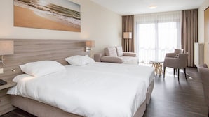 Habitación de hotel con una cama grande, dos mesitas de noche, un escritorio, una silla, una pequeña mesa redonda y un cuadro con una playa.