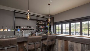 Un espace bar avec des tabourets, un comptoir et une étagère remplie de bouteilles. On y trouve également des luminaires suspendus et une fenêtre donnant sur un beau paysage.