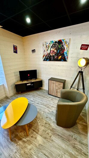 A room with a wooden floor, a TV on a wooden stand, a colorful abstract painting on the wall, a tripod lamp, and a brown armchair. - Family Apartment in La Bourboule (La Bourboule)