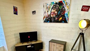 Un soggiorno moderno con una TV a schermo piatto, un quadro astratto colorato, una lampada da terra con treppiede e un tavolo giallo e grigio dal design originale.
