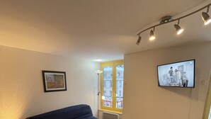 A living room with a dark blue sofa, a wooden coffee table, a small side table with a lamp, a TV mounted on the wall, and a window with a view of buildings. - Bright Apartment in Mont-dore (Mont-Dore)