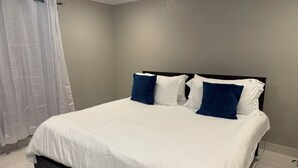 Un letto rifatto con lenzuola bianche e cuscini blu, una testiera nera e una tenda bianca su una parete di colore chiaro.