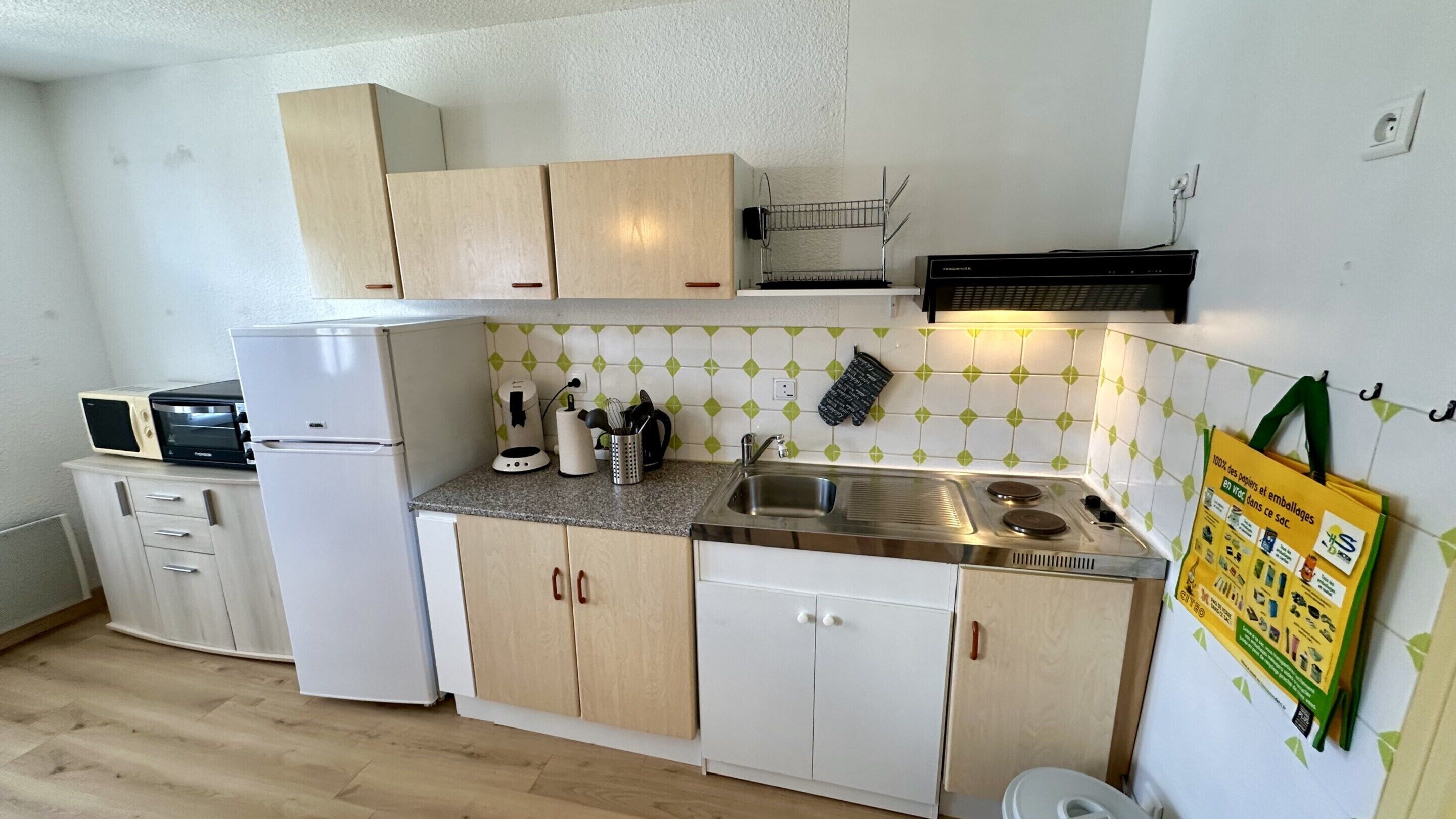 Een compacte keuken met een koelkast, magnetron, gootsteen en fornuis. Er zijn kastjes en een klein aanrecht.