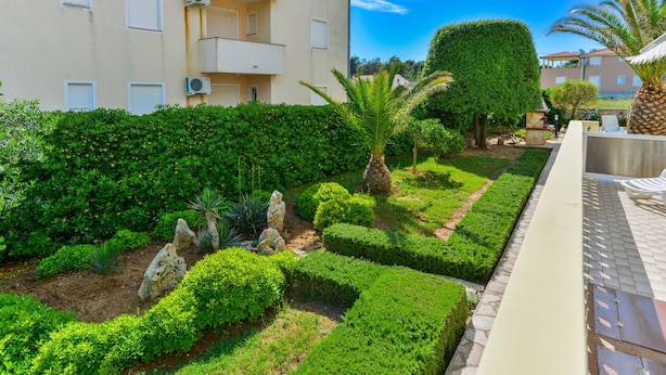 A well-maintained garden with neatly trimmed hedges, a palm tree, and a stone sculpture. - Beachfront Apartment for 4 (Vir)
