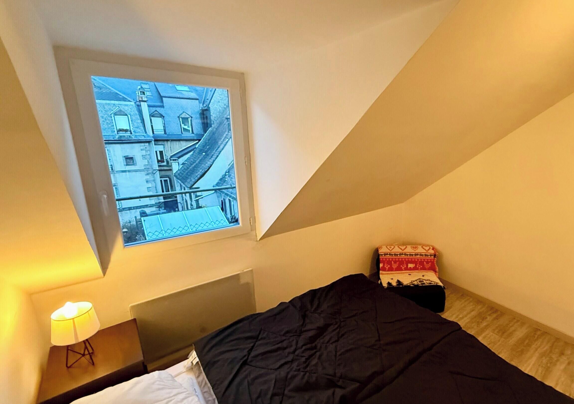 A bedroom with a bed, a nightstand with a lamp, a window with a view of buildings, and a small bedside table with a patterned pillow.