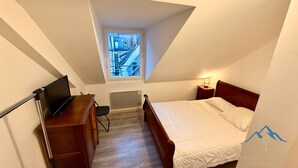 Pequeña habitación triangular con una cama, un escritorio y una silla. Se ve una ventana con vistas a edificios.
