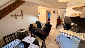 Un espacio de sala cómodo con un sofá negro, una mesa de comedor con manteles estampados, una cafetera y un refrigerador pequeño.