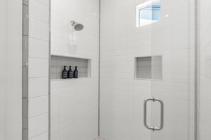 A modern shower area with white tiles, a handheld showerhead, and a small shelf with bottles.