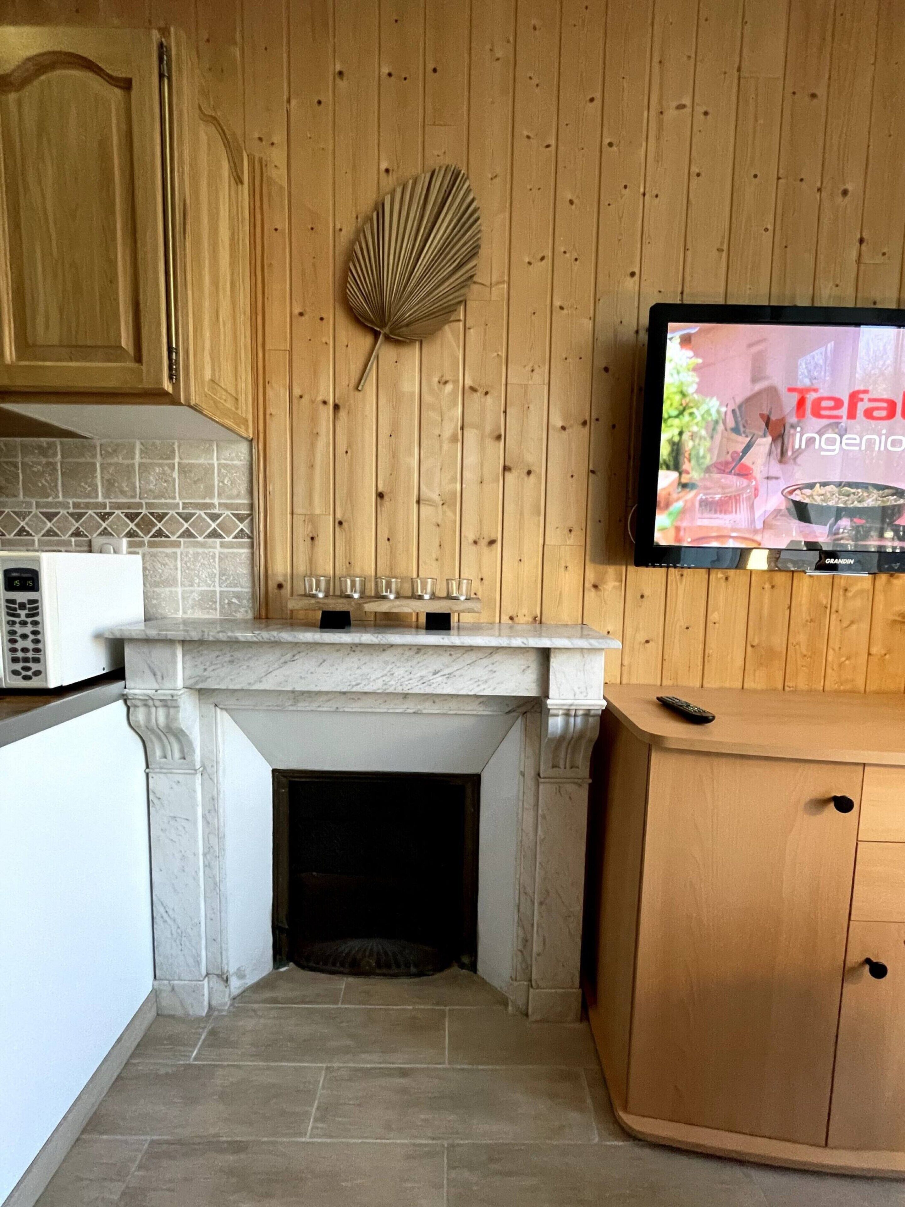 A kitchen with wooden cabinets, a tiled backsplash, a marble fireplace, and a television mounted on the wall.