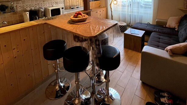 Una cocina moderna con armarios de madera, una isla central con taburetes, un televisor y vistas a la calle a través de grandes ventanales.
