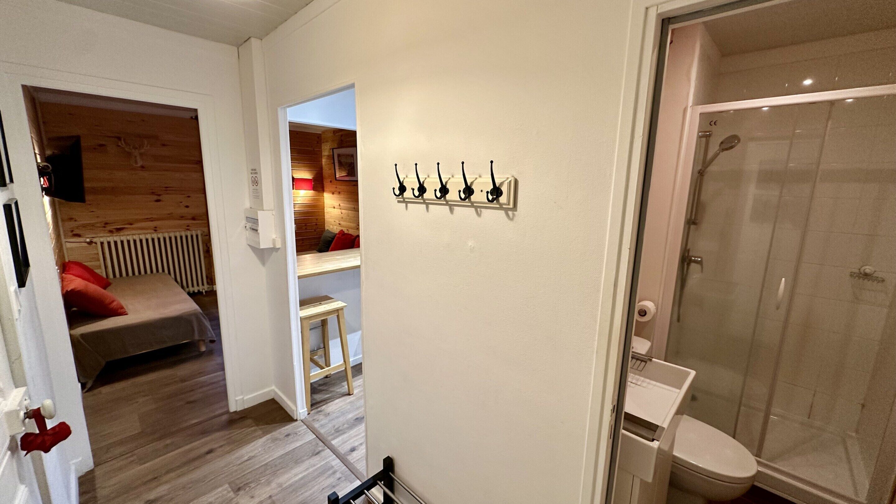 Habitación de hotel con piso de madera, cama con almohadas rojas, un taburete pequeño, un panel de ganchos en la pared y zona de ducha.
