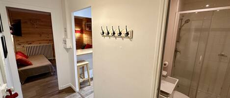 Habitación de hotel con piso de madera, cama con almohadas rojas, un taburete pequeño, un panel de ganchos en la pared y zona de ducha.
