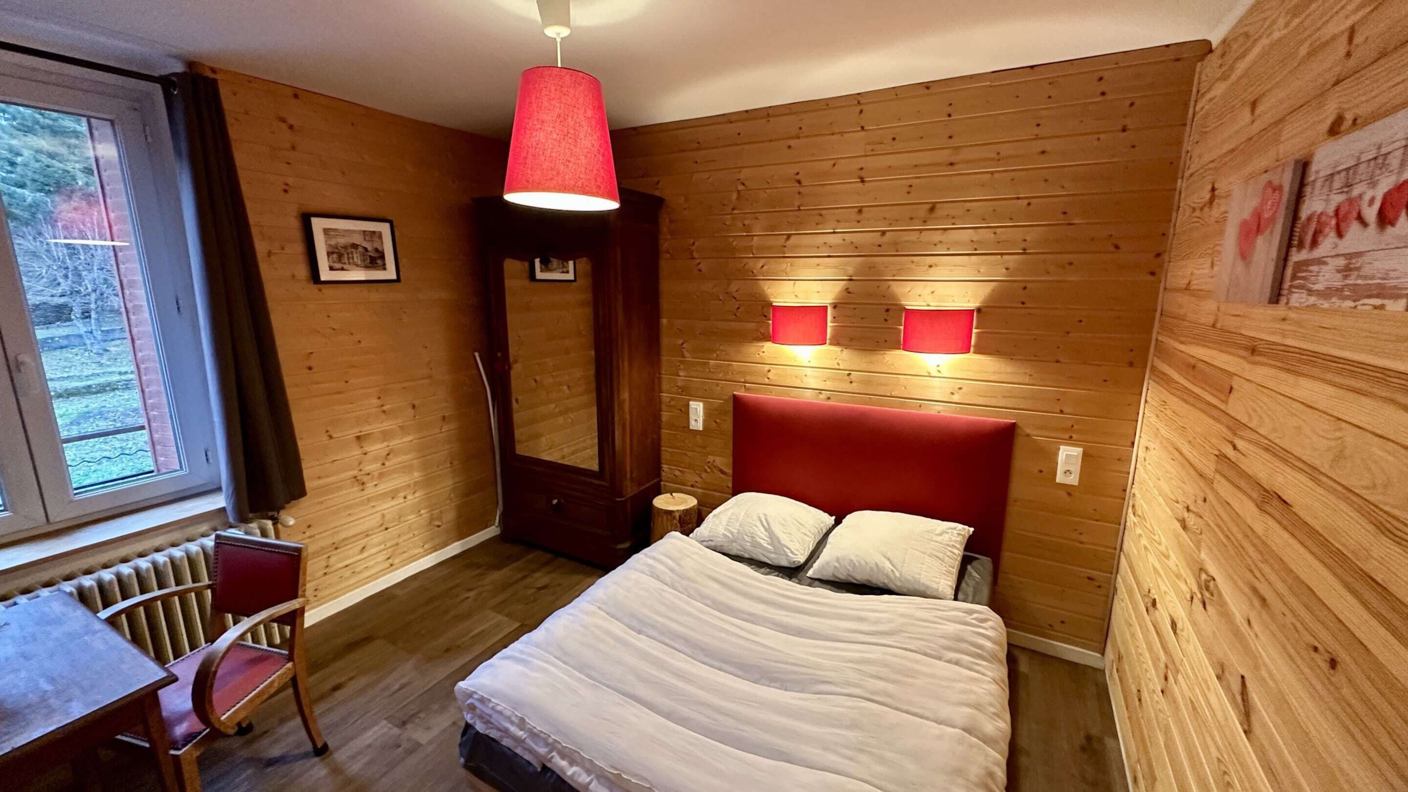Une pièce aux murs lambrés, comprenant un lit, un bureau et deux lampes fixées au mur.