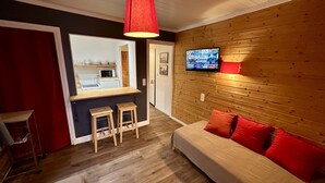 Un espacio habitable compacto con kitchenette, un sofá con cojines rojos, un televisor montado en la pared y piso de madera.