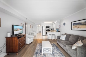 Ein modernes Wohnzimmer mit Fernseher, Sofa und Couchtisch. Im Hintergrund ist eine Küche mit weißen Schränken und einer Mikrowelle zu sehen.