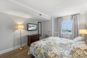 Ein Hotelzimmer mit Bett, einem an der Wand befestigten Fernseher, einem Schminktisch, einer Lampe und einem Fenster mit Vorhängen.