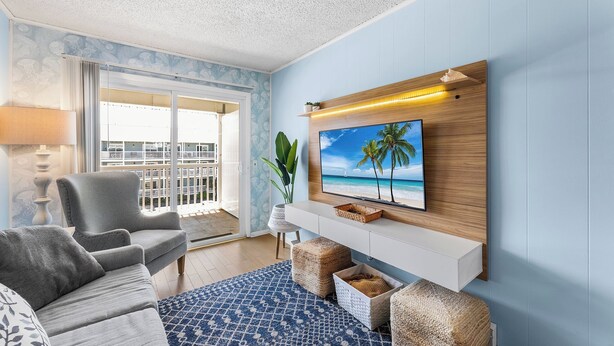 Ein Wohnzimmer mit grauem Sofa, einem blauen Teppich mit Muster, einem Fernseher an einer Holzwand und einem Balkon mit Blick.
