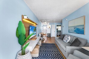 A modern living room with a sofa, a dining area, and a kitchen in the background. - Atlantic Court 303 Steps to the beach (Ocean City)