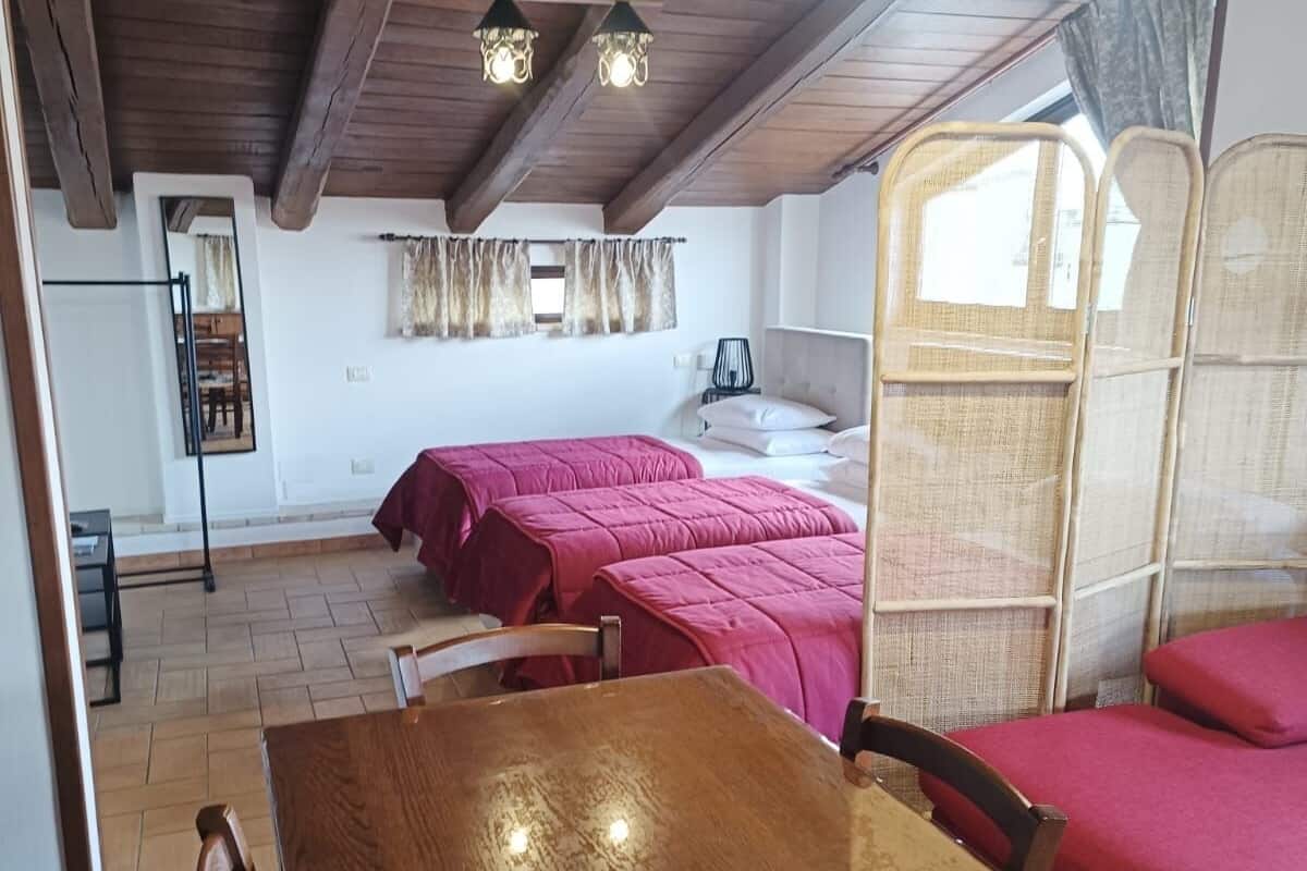 A room with wooden beams, a wooden table, two beds with red bedding, a mirror, and a window with curtains.