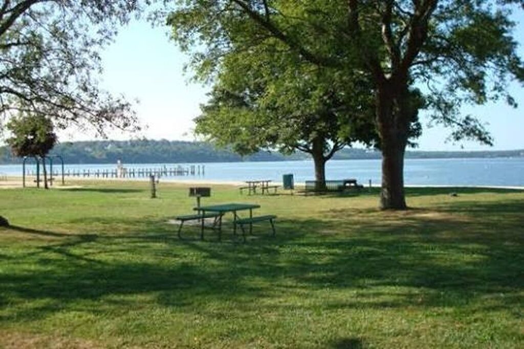 A grassy park area with picnic tables and benches, a large tree, and a view of a body of water.