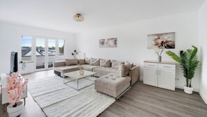 Una sala de estar moderna con un sofá modular, una mesa de centro y una alfombra blanca. Hay un televisor a la izquierda, un mueble a la derecha y una planta en la esquina.