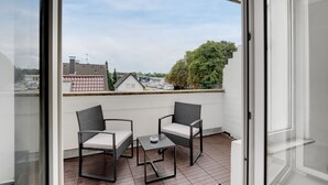 Un balcón con dos sillas de mimbre y una mesita, con vistas a una zona residencial con casas y árboles.