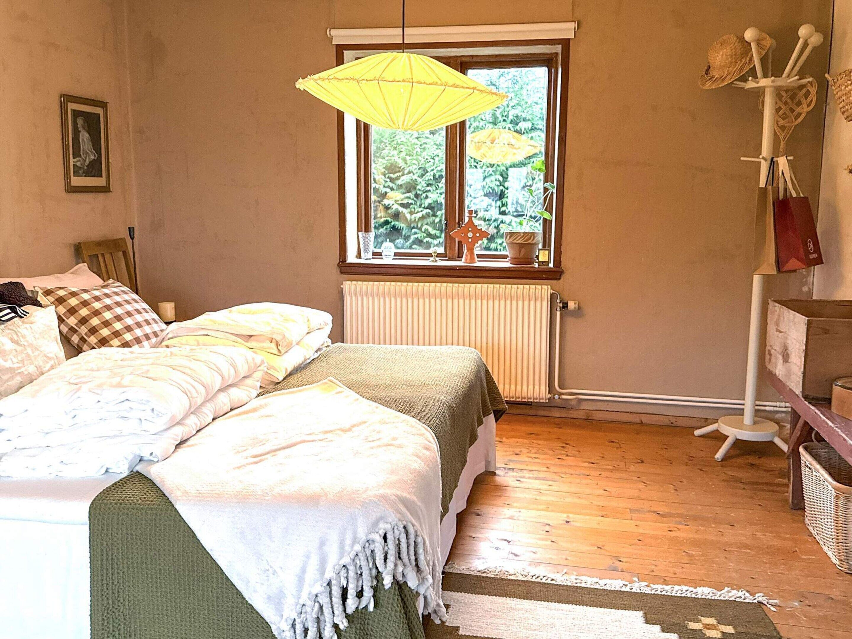 A bedroom with a bed, a window with a view of greenery, a radiator, and a shelf with items.
