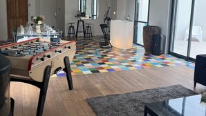 A modern living room with a foosball table, a coffee table, and a sofa. There is a dining area with a table set for four and a bar area with stools. The room has large windows and a colorful patterned floor. - Near La Baule/Historical center.  Villa located in a bubble of Serenity. (Guérande)