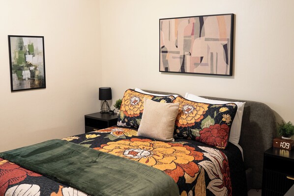 A neatly arranged bedroom with a floral bedspread, a modern abstract painting, and a digital clock on a bedside table.
