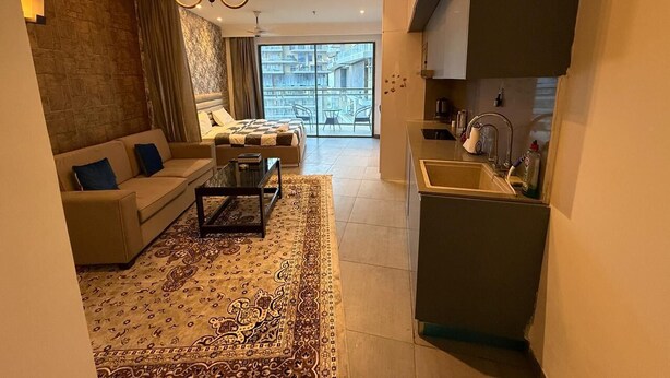 A compact living space with a sofa, coffee table, and patterned rug. A kitchen area with a sink and cabinets is visible. - spira.staycation (Noida)