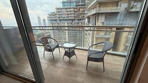 A balcony with wicker chairs and a glass table, overlooking a high-rise building with construction cranes. - spira.staycation (Noida)