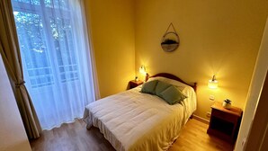 A bedroom with a bed, two bedside tables, a wall-mounted lamp, and a window with curtains. - So' Amandine - La Bourboule (La Bourboule)