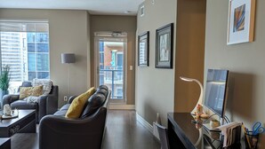 Uma sala de estar moderna com um sofá de couro escuro, uma mesa de centro e uma escrivaninha com um computador e abajur. Há quadros nas paredes e uma janela com persianas.