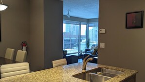 Cozinha moderna com bancadas de granito, pia de aço inoxidável e lava-louças embutida. Há um espaço de jantar com mesa e cadeiras, e uma janela grande que oferece vista da cidade.