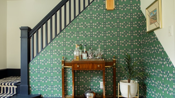Una escalera con barandilla azul oscuro, pared de diseño verde, consola de madera con botellas y una planta en maceta, y alfombra a rayas.