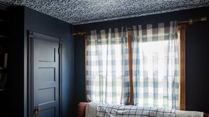 Una habitación con una pared azul oscuro, techo estampado, una ventana con cortinas a cuadros, una puerta y un sofá con una manta a cuadros.