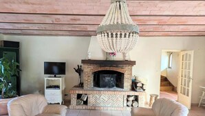 Una sala de estar con chimenea, dos sillones blancos, una mesa de centro de madera y un televisor sobre un mueble blanco.