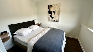 A neatly made bed with white linens and a gray throw blanket, a bedside table with a lamp, and a wall-mounted abstract painting. - Les Etrangers 402 - La Bourboule (La Bourboule)