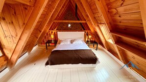 A high-ceilinged wooden room with a bed centered, two bedside tables with lamps, and a small window at the far end.