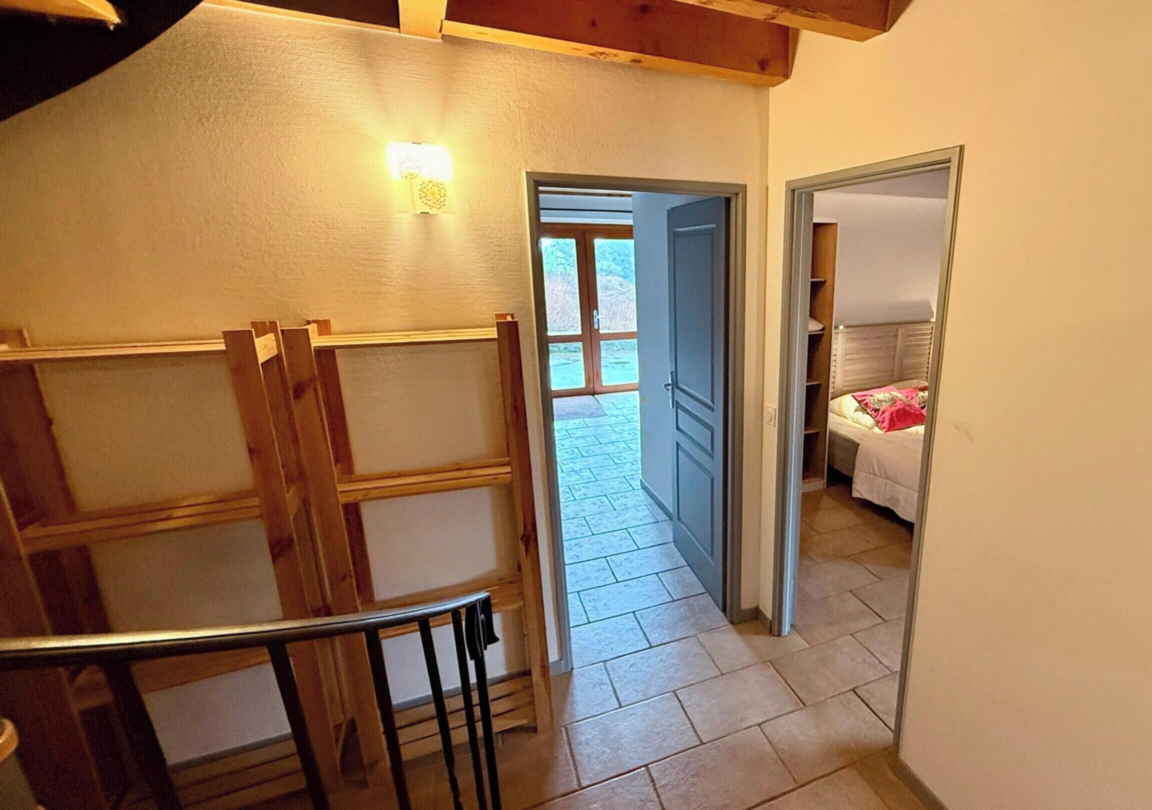Corredor estreito com escada de madeira à esquerda, porta que dá para um quarto com cama e espelho, e vista para o exterior pela porta.