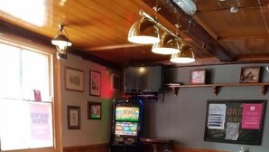 Une table de billard se trouve dans une pièce aux murs en bois et au plafond bas. On aperçoit, à l’arrière-plan, des tabourets et une borne d’arcade illuminée par des néons.