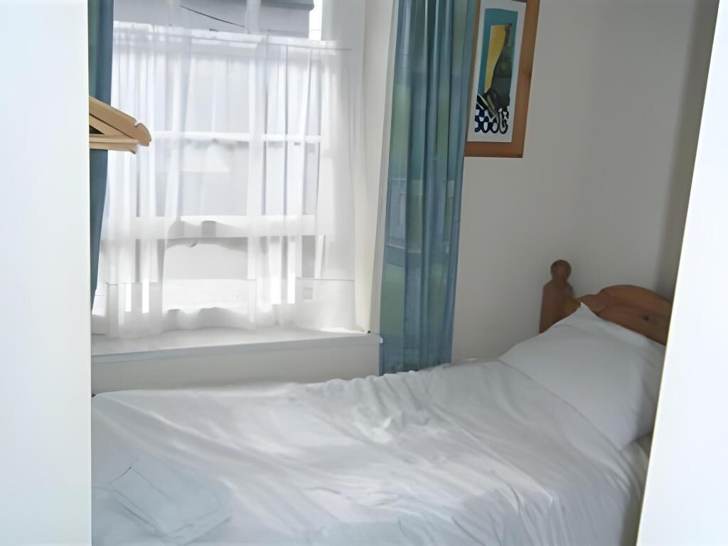 A single bed with white bedding in a room with a window, a wooden headboard, and a framed picture on the wall.