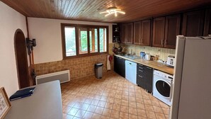 A kitchen with wooden cabinets, a sink, a washing machine, and a window with a view of greenery. - Villa Janchrisma - Montdore - 11pers (Mont-Dore)