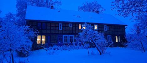A two-story house with a snow-covered roof and windows, surrounded by snow-laden trees.