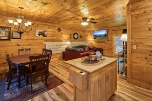 A wooden cabin interior with a dining table, chairs, a sofa, a TV, and a kitchen area. - Private cozy log cabin-Your sweet spot in Smoky (Pigeon Forge)