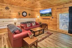 A cozy cabin interior with a red leather sofa, a wooden coffee table, a mounted TV, and a window with a view of a snowy landscape. - Private cozy log cabin-Your sweet spot in Smoky (Pigeon Forge)
