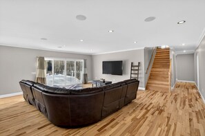 A modern living room with a sectional sofa, a flat-screen TV, and a wooden staircase. - Solar-Powered Luxury Estate | Master Spa, Tesla EV, Fast WiFi & Game Room! (Tolland)