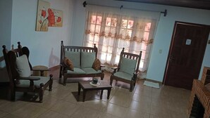 Una sala de estar con techo de madera, un ventanal con cortinas translúcidas, un cuadro en la pared y una puerta a la derecha.