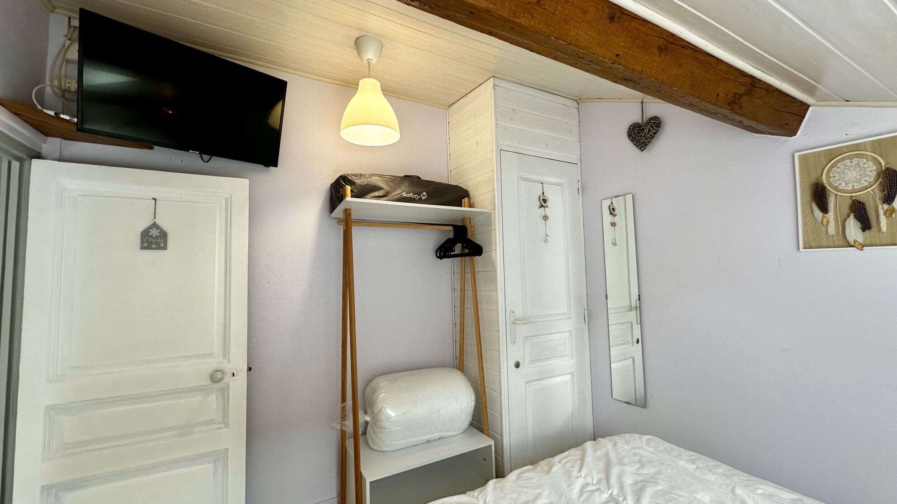 A bedroom with a bed, a wardrobe, a mirror, a hanging light fixture, and a television mounted on the wall.